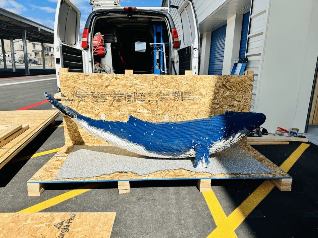 Large, artistic whale sculpture made of wooden crates, positioned on a pallet outside a delivery van, showcasing creative packaging solutions related to international shipping.