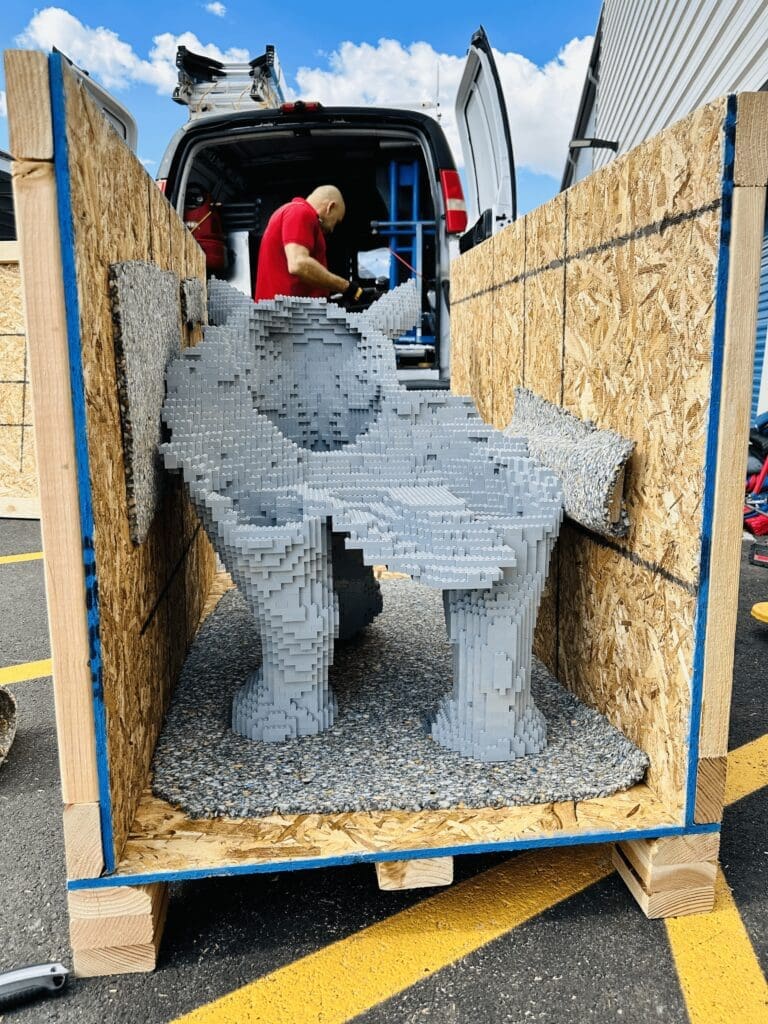 Custom wooden crate containing a sculptural piece made of gray blocks, with a worker in a red shirt preparing the crate for shipping, highlighting the importance of sturdy packaging for fragile items.