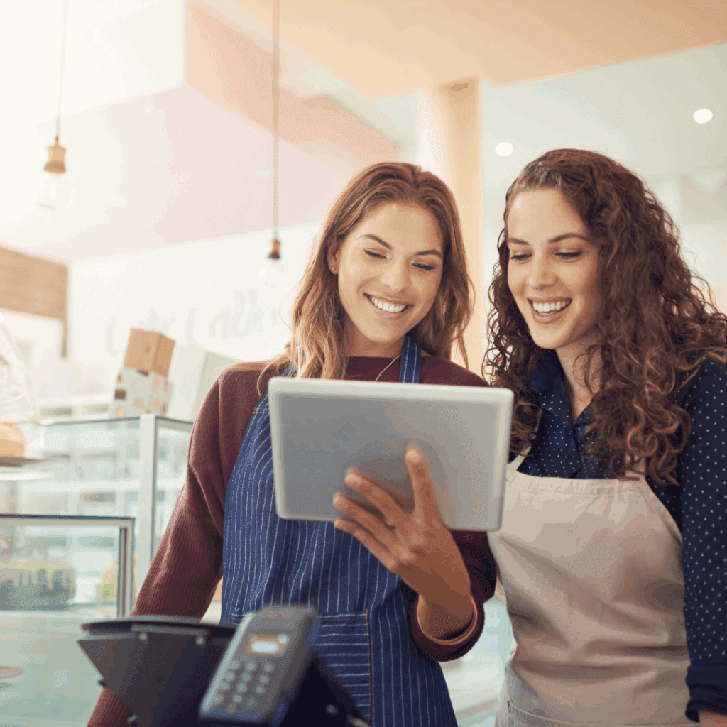 Two women in aprons smiling and looking at a tablet in a modern retail setting, reflecting teamwork and customer service in a vibrant environment.